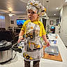 apron, barefoot, brown_pants, ceiling_lamp, chef_hat, child, countertop, curtains, fan, indoor, kitchen, living_room, person, plant, plastic_container, pressure_cooker, television, toddler, wooden_cutting_board, yellow_shirt