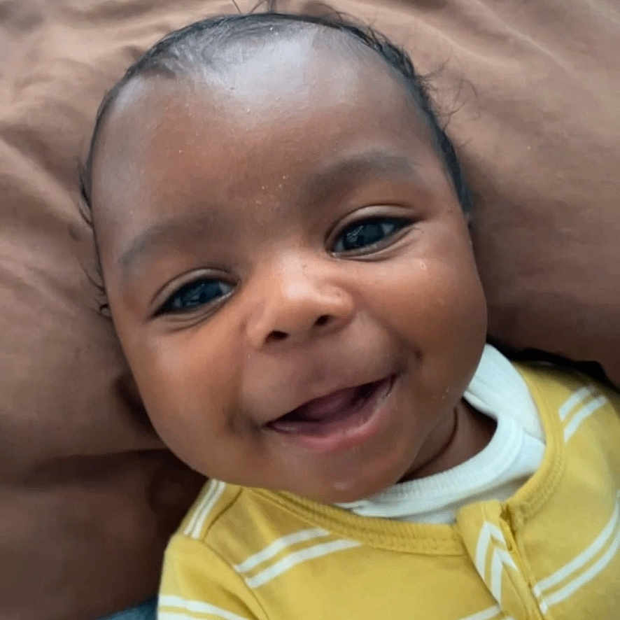 Kyvie is registered to the contest to win money with this photo: baby, child, closeup, cute, eyes, face, happy, head, indoor, infant, mouth, person, pillow, portrait, resting, skin, smiling, striped, yellow_clothing, young_child