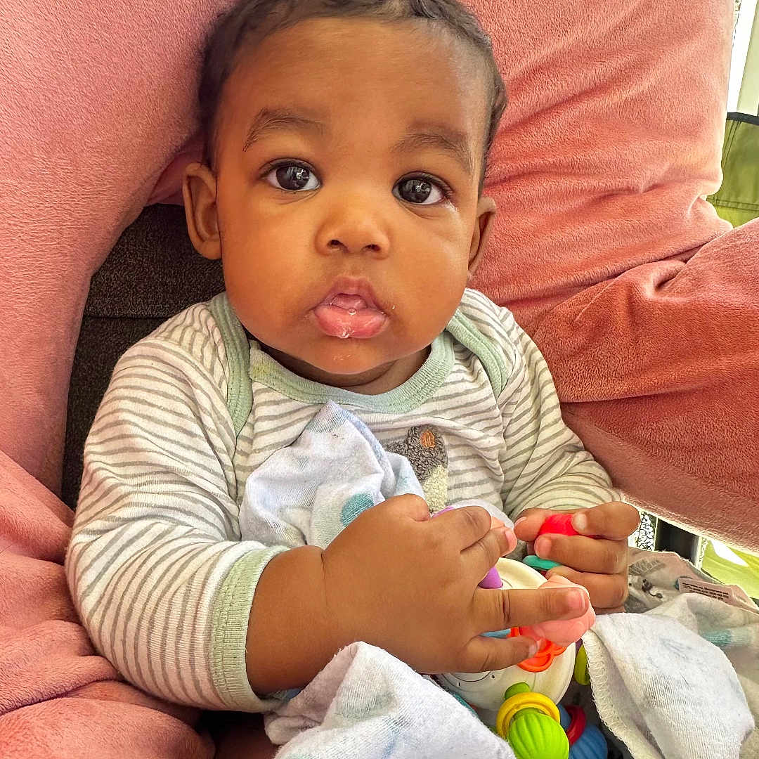 Taevon is registered to the contest to win money with this photo: baby, child, infant, toy, blanket, striped_clothing, pink_chair, indoor, cute, curious, sitting, face, hands, expression, plush, comfort, young_child, portrait, cozy, colorful