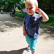 Aloïs a rejoint le concours — aidez-le/la à gagner de superbes lots ! adult, bench, blue_shorts, casual_clothing, child, daytime, greenery, happy, leaf_pattern, outdoor, park, pathway, playful, sandals, shadow, smiling, sunlight, toddler, trees, wristband