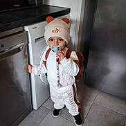 Leyann a rejoint le concours — aidez-le/la à gagner de superbes lots ! toddler, child, pacifier, hat, bear_ears, backpack, kitchen, dishwasher, floor, tile, white_clothing, brown_clothing, black_shoes, standing, indoor, appliance, cute, person, home, flooring