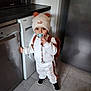 toddler, child, pacifier, hat, bear_ears, backpack, kitchen, dishwasher, floor, tile, white_clothing, brown_clothing, black_shoes, standing, indoor, appliance, cute, person, home, flooring