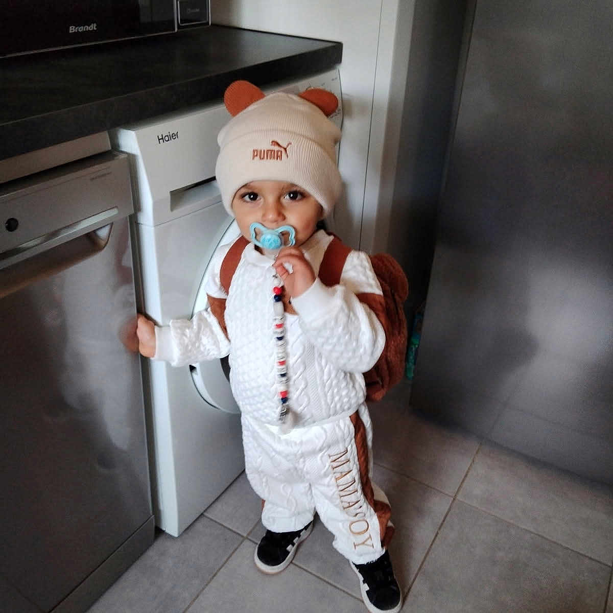 Leyann a rejoint le concours — aidez-le/la à gagner de superbes lots ! appliance, backpack, bear_ears, black_shoes, brown_clothing, child, cute, dishwasher, floor, flooring, hat, home, indoor, kitchen, pacifier, person, standing, tile, toddler, white_clothing