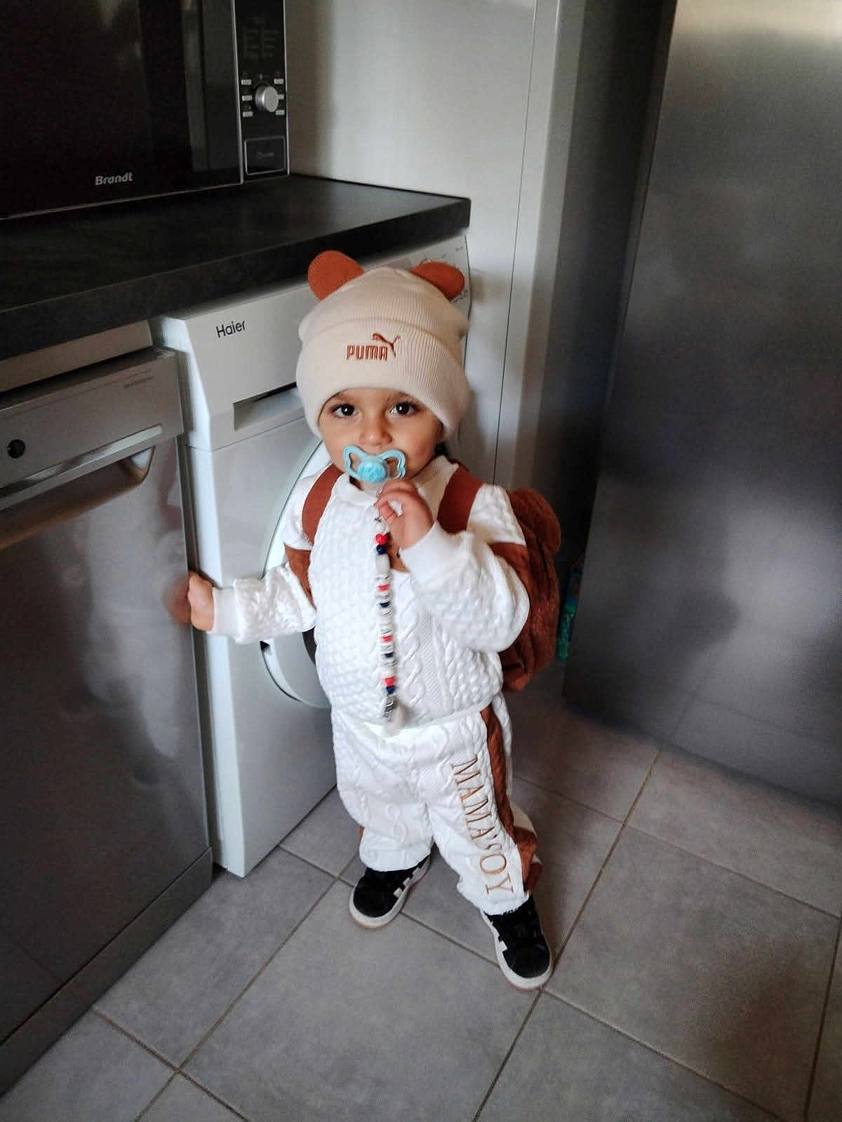 Leyann a rejoint le concours — aidez-le/la à gagner de superbes lots ! toddler, child, pacifier, hat, bear_ears, backpack, kitchen, dishwasher, floor, tile, white_clothing, brown_clothing, black_shoes, standing, indoor, appliance, cute, person, home, flooring