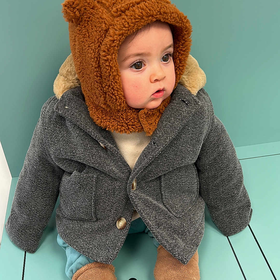 Noah Lorenzo a rejoint le concours — aidez-le/la à gagner de superbes lots ! baby, bear_ears, brown_hat, child, coat, curious, cute, face, footwear, fuzzy, gray_coat, hat, indoor, portrait, sitting, socks, teal_bench, warm_clothing, winter_wear, young_child