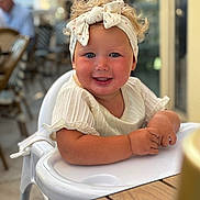 Aleya participe au concours pour gagner de l'argent avec cette photo : toddler, child, high_chair, smiling, headband, curly_hair, blush, white_clothing, table, indoor, cafe, person, portrait, happy, cute, baby, seat, furniture, skin, face