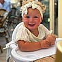 toddler, child, high_chair, smiling, headband, curly_hair, blush, white_clothing, table, indoor, cafe, person, portrait, happy, cute, baby, seat, furniture, skin, face
