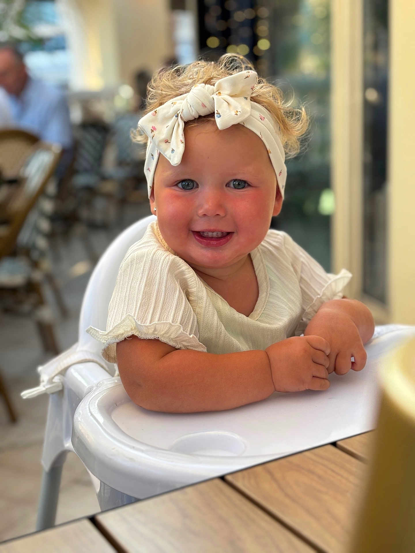 Aleya participe au concours pour gagner de l'argent avec cette photo : toddler, child, high_chair, smiling, headband, curly_hair, blush, white_clothing, table, indoor, cafe, person, portrait, happy, cute, baby, seat, furniture, skin, face