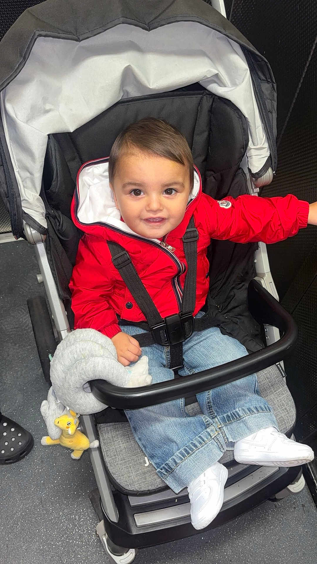Kayaan is registered to the contest to win money with this photo: baby, black_stroller, casual_clothing, child, cute, floor, happy, indoor, jeans, person, playful, portrait, red_jacket, seatbelt, smile, stroller, toddler, toy, white_shoes, young_child