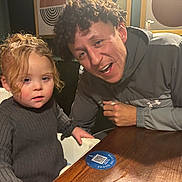 Danziell is registered to the contest to win money with this photo: child, man, curly_hair, high_chair, table, indoor, sweater, cup, wooden_table, person, face, seated, curly_hair_child, curly_hair_man, cozy, casual_clothing, warm_lighting, restaurant, QR_code, interaction