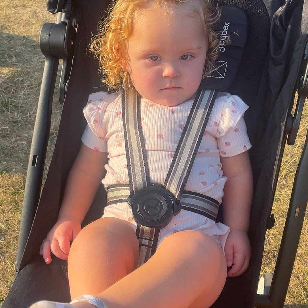 Danziell is registered to the contest to win money with this photo: baby, black_stroller, child, curly_hair, daylight, grass, legs, outdoor, park, person, picnic_table, pink_clothing, plaid_pattern, portrait, seatbelt, serious_expression, socks, stroller, sunlight, toddler