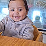 toddler, child, smiling, hoodie, drool, high_chair, wooden_table, indoor, face, happy, dimples, clothing, person, baby, seat, table, casual, furniture, young_child, portrait