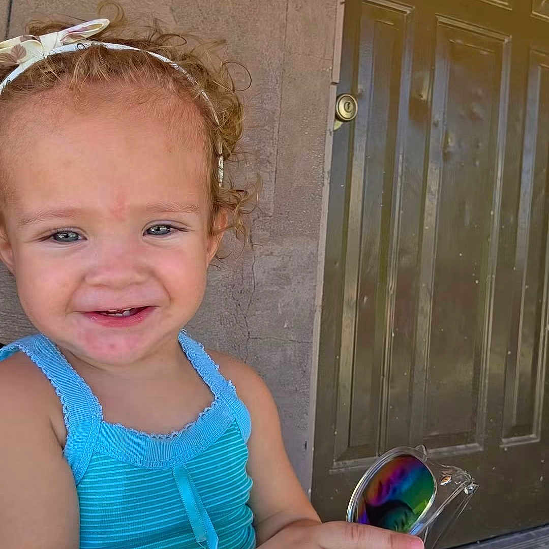 Maria is registered to the contest to win money with this photo: child, toddler, smiling, portrait, outdoor, doorway, front_door, wall, curly_hair, hair_bow, blue_clothing, tank_top, sunglasses, rainbow_lenses, hand, fashion, happy, face, standing, daylight
