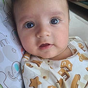 Christopher is registered to the contest to win money with this photo: baby, bed, bedding, child, close_up, clothing, dark_hair, face, home, indoors, infant, looking_at_camera, lying_down, nursery, onesie, patterned_clothing, portrait, sitting, soft_lighting, wide_eyes