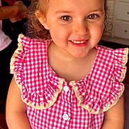 Promiss is registered to the contest to win money with this photo: child, girl, smile, crown, pink, dress, checkered, curly_hair, table, indoor, kitchen, person, button, happy, portrait, celebration, cute, young, hands, face
