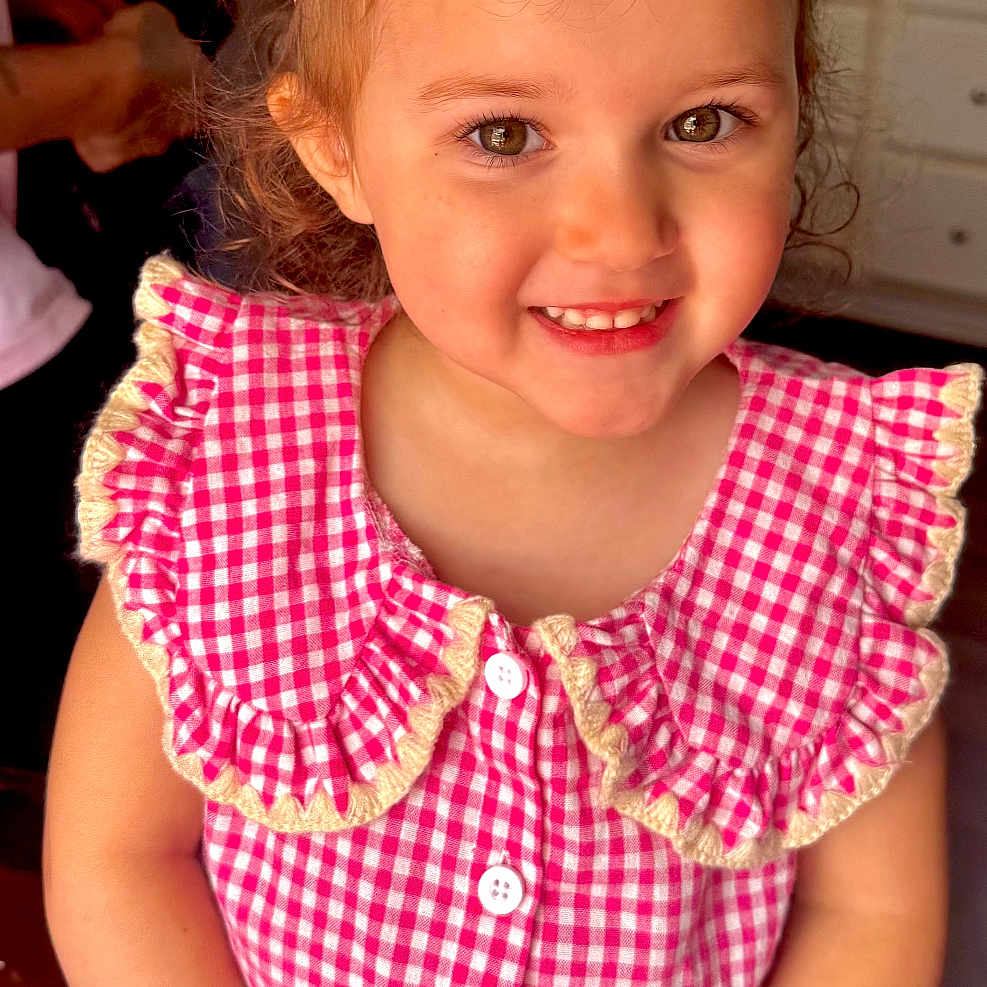 Promiss is registered to the contest to win money with this photo: child, girl, smile, crown, pink, dress, checkered, curly_hair, table, indoor, kitchen, person, button, happy, portrait, celebration, cute, young, hands, face