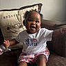 baby, child, smiling, sitting, couch, portrait, indoor, home, cushion, toy, bracelet, hair_bows, curly_hair, white_shirt, colorful_clothing, floral_shorts, socks, happy, face, photography