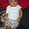 baby, child, infant, portrait, lying_down, bed, indoor, bow_headband, checkerboard, white_shirt, patterned_shorts, socks, dark_skin, face, eyes, serious_expression, fashion, textile, bedding, photography