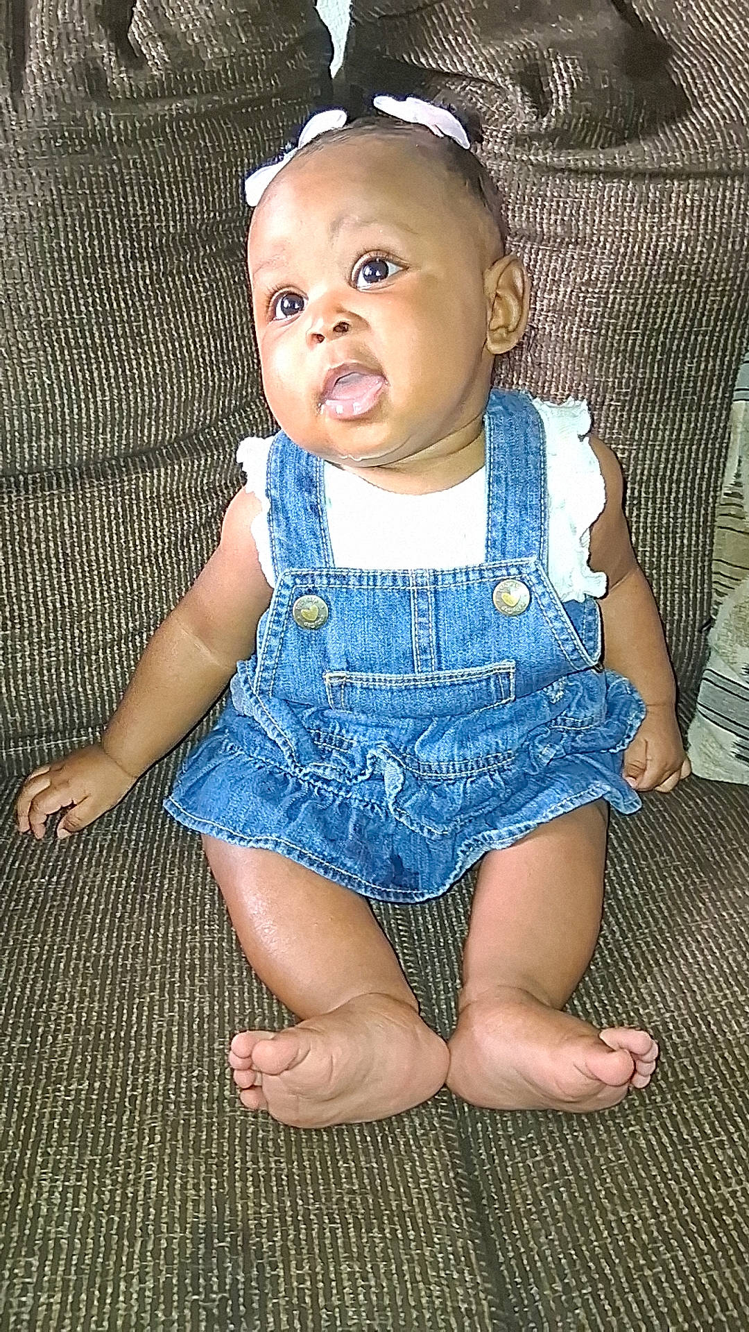 baby, bow, child, clothing, couch, denim, dress, eyes, face, feet, furniture, home, indoor, infant, overalls, photography, portrait, sitting, smiling, toes