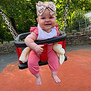 Willow is registered to the contest to win money with this photo: baby, barefoot, blanket, bow, chains, child, greenery, happy, headband, infant, outdoor, park, pink_clothing, playground, seat, smiling, stone_wall, sunlight, swing, trees