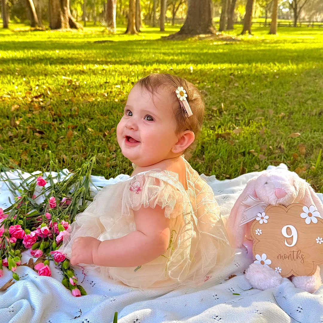 Amelia joined the competition — help win amazing prizes! baby, child, infant, girl, dress, blanket, flowers, grass, park, outdoor, sunlight, plush_toy, bunny, sign, nature, smiling, happy, portrait, sitting, cute
