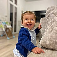 Swann participe au concours pour gagner de l'argent avec cette photo : toddler, child, smiling, indoor, couch, blue_sweater, white_shirt, hair_clip, happy, standing, furniture, living_room, flooring, window, toy, baby, person, home, cute, portrait