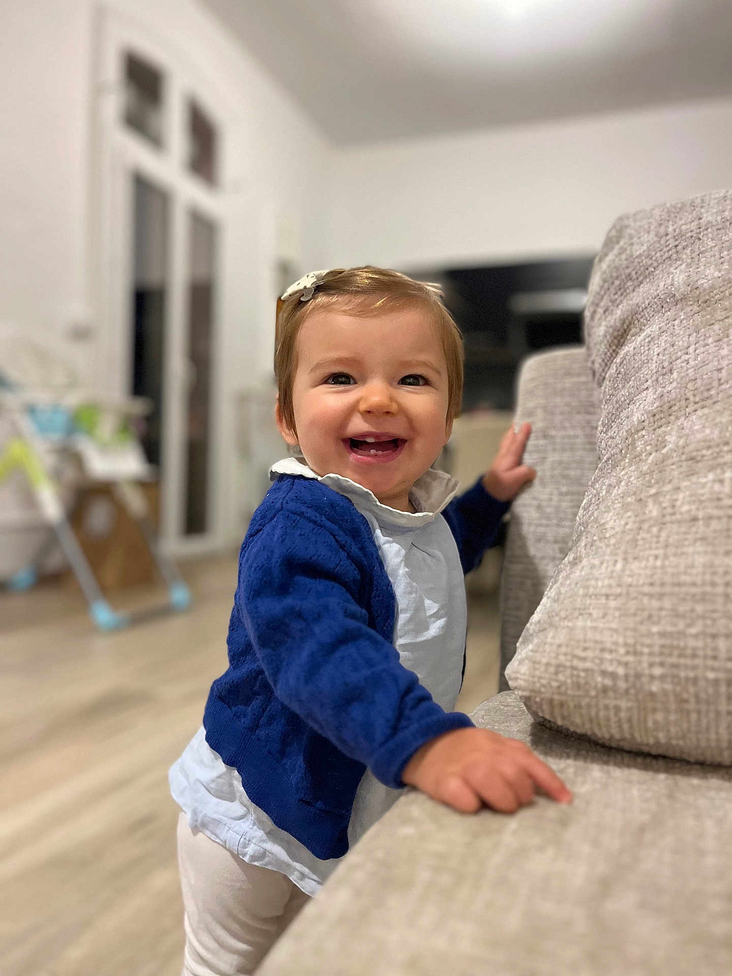 Swann participe au concours pour gagner de l'argent avec cette photo : toddler, child, smiling, indoor, couch, blue_sweater, white_shirt, hair_clip, happy, standing, furniture, living_room, flooring, window, toy, baby, person, home, cute, portrait