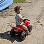 Isaac is registered to the contest to win money with this photo: toddler, child, toy, atv, red, outdoor, dirt, path, mobile_home, blue, grass, sunny, daylight, curly_hair, smile, casual_clothing, footwear, recreational_vehicle, rural, play