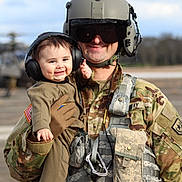 Benaiah is registered to the contest to win money with this photo: soldier, baby, military_uniform, headphones, smile, outdoor, helicopter, camouflage, helmet, gloves, toddler, person, portrait, army, airfield, equipment, happy, holding, face, protection