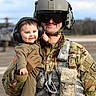 soldier, baby, military_uniform, headphones, smile, outdoor, helicopter, camouflage, helmet, gloves, toddler, person, portrait, army, airfield, equipment, happy, holding, face, protection