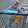 toddler, child, shopping_cart, pacifier, knit_hat, sweater, plush_toy, sneakers, pants, outdoor, street, crosswalk, curious, baby, clothing, person, toy, casual, daylight, urban
