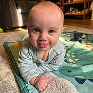 Patrick is registered to the contest to win money with this photo: baby, blanket, blue_eyes, books, carpet, child, couch, cozy, cute, dinosaur_pattern, floor, furniture, home, indoor, infant, pajamas, play_mat, smiling, tongue_out, wooden_shelf