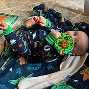 Patrick is registered to the contest to win money with this photo: baby, infant, toy, blanket, pajamas, dinosaur_pattern, star_wars, floor, wood_floor, carpet, foot, hand, face, indoor, child, play, colorful, cute, relaxed, lying_down