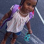 child, girl, braids, beads, bow, backpack, glasses, parking_lot, asphalt, sandals, shorts, striped_top, smiling, standing, outdoor, person, jewelry, accessories, casual_wear, daytime