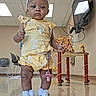 toddler, child, yellow_outfit, headband, bow, shoes, indoor, floor, activity_table, television, chair, ceiling, lights, reflection, person, baby, expression, standing, playroom, toy