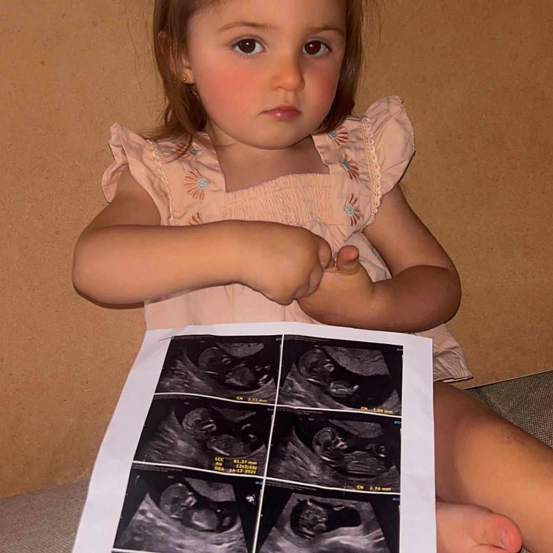 Téa participe au concours pour gagner de l'argent avec cette photo : baby_images, barefoot, child, closeup, couch, face, hair_tied, holding, indoor, medical_images, pacifier, person, pink_clothing, portrait, printout, serious_expression, sitting, toddler, ultrasound, young_child