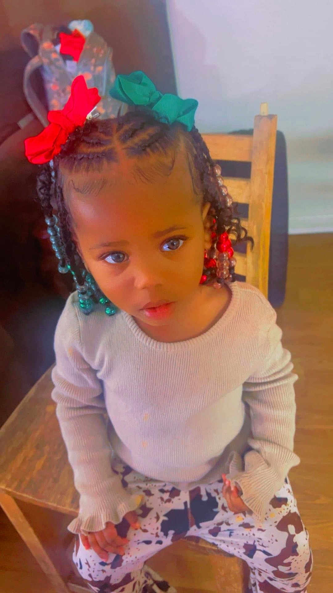 Taziyah joined the competition — help win amazing prizes! child, girl, braided_hair, hair_bows, red_bow, green_bow, sitting, wooden_chair, beige_sweater, patterned_pants, indoor, backpack, curious_expression, young_child, person, cute, fashion, portrait, home, floor