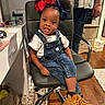 toddler, child, indoor, chair, denim_overalls, boots, hair_bows, person, floor, rug, cabinet, hand, hair, smile, white_tshirt, jeans, home, furniture, adult, accessory