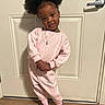 child, girl, smile, pose, pink_clothing, tracksuit, nike_logo, hair_puffs, brown_slippers, door, wooden_floor, indoors, casual_wear, necklace, happy, cute, standing, portrait, young_child, face