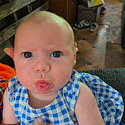 Samantha is registered to the contest to win money with this photo: baby, infant, face, blue_eyes, dress, checkered_pattern, food, mouth, expression, indoor, floor, wood, furniture, person, child, cute, skin, hand, sitting, closeup