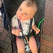 Arthur a rejoint le concours — aidez-le/la à gagner de superbes lots ! baby, stroller, child, blue_eyes, holding, pink_straw, green_straw, white_shirt, sitting, curious, outdoor, pavement, black_stroller, speckled_pattern, barefoot, summer_clothing, cute, infant, seatbelt, toy