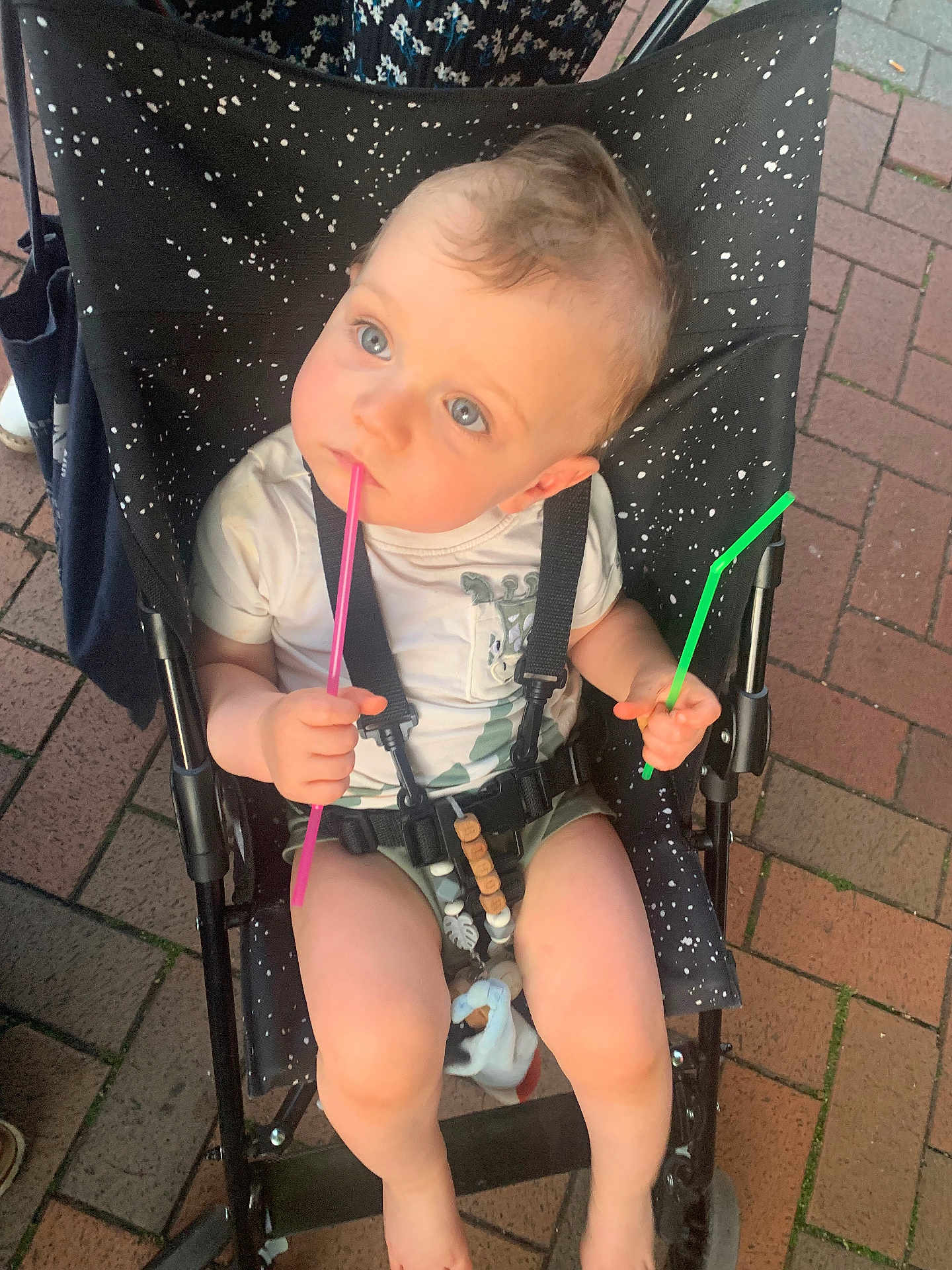 Arthur a rejoint le concours — aidez-le/la à gagner de superbes lots ! baby, stroller, child, blue_eyes, holding, pink_straw, green_straw, white_shirt, sitting, curious, outdoor, pavement, black_stroller, speckled_pattern, barefoot, summer_clothing, cute, infant, seatbelt, toy