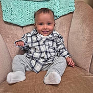 Zakariya is registered to the contest to win money with this photo: baby, child, smiling, plaid_shirt, gray_pants, socks, armchair, knitted_blanket, beige, cozy, indoor, portrait, happy, person, cute, seated, home, infant, clothing, furniture