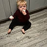 child, toddler, blond_hair, sitting, floor, indoor, portrait, face, smiling, snacking, food, treat, hand, barefoot, dark_clothing, red_shirt, black_pants, cabinets, wood_floor, candid