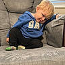 child, boy, toddler, blond_hair, sleepy, sitting, recliner, armchair, blue_sweatshirt, black_pants, colorful_socks, indoors, living_room, window, sunlight, portrait, relaxed, cozy, furniture, home