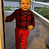 child, toddler, boy, portrait, standing, doorway, outdoor, building, architecture, red_pants, plaid_shirt, black_boots, blond_hair, smiling, face, full_body, sunlight, railing, sidewalk, urban