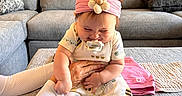 Valentina is registered to the contest to win money with this photo: baby, child, pacifier, headband, flower, bee_pattern, sitting, couch, gray_couch, living_room, hands, supporting, cute_outfit, adult, ottoman, cozy, indoor, smiling, happy, home