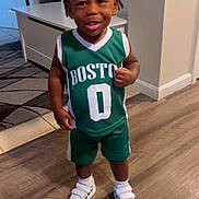Jeremiah is registered to the contest to win money with this photo: toddler, child, smiling, indoors, basketball_jersey, green_clothing, shorts, white_shoes, socks, wooden_floor, table, rug, person, happy, standing, hair, face, expression, home, flooring