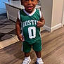 toddler, child, smiling, indoors, basketball_jersey, green_clothing, shorts, white_shoes, socks, wooden_floor, table, rug, person, happy, standing, hair, face, expression, home, flooring