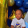 child, toddler, balloons, birthday, number_one_balloon, gold_balloon, blue_balloon, sneakers, white_tshirt, curly_hair, indoor, celebration, party, couch, seated, holding_shoe, young_child, portrait, cute, happy